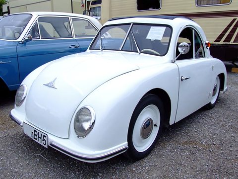 White rounded small coupé on gravel beside a blue saloon, a caravan behind. Opens a larger view