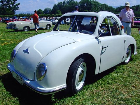 White rounded small coupé with plate BH 5 on sunny grass, visitors and other classic cars behind. Opens a larger view