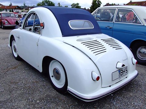 White small car with a navy fabric roof and louvred tail on gravel, a blue saloon beside it. Opens a larger view