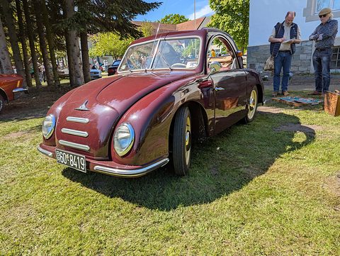 Maroon small car with a chrome-striped nose on sunny grass, two men looking at papers beside a wall. Opens a larger view