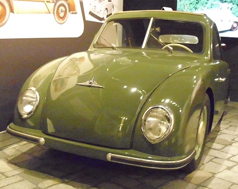 Olive green rounded small car on a cobbled display floor, factory artwork on the wall behind. Opens a larger view