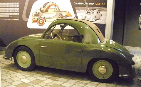 Olive green small car with its fabric roof folded, a large cutaway drawing and “Champion 400 1951-52” sign behind. Opens a larger view