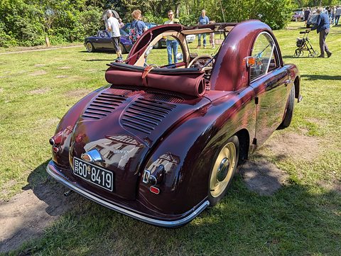 Maroon small car from behind on grass, louvred tail and a rolled roof, visitors beyond. Opens a larger view