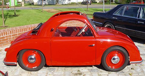 Bright red small convertible saloon with red wheels on cobbles beside a dark limousine. Opens a larger view