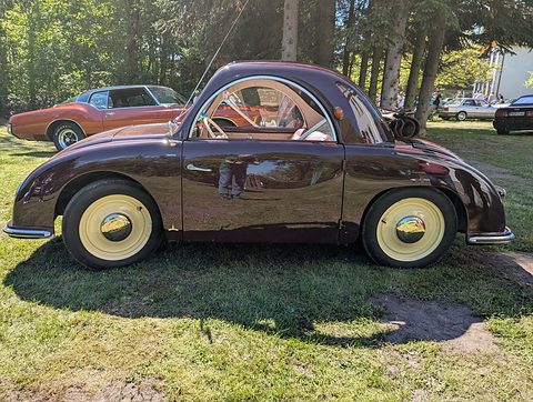 Maroon small car with cream wheels in side view on grass under pine trees. Opens a larger view