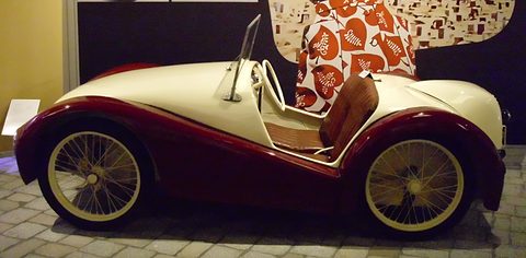Side view of a cream and maroon doorless two-seater with a brown seat and wire wheels. Opens a larger view