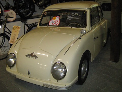 Cream small two-door saloon with a winged Maico badge, bicycles on the wall behind.