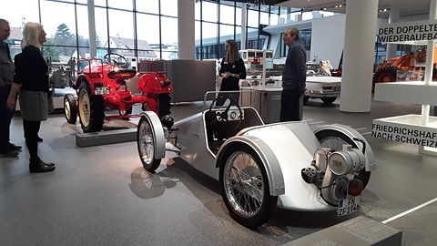 Silver roadster with an exposed rear engine on a grey floor, visitors and a red tractor behind. Opens a larger view