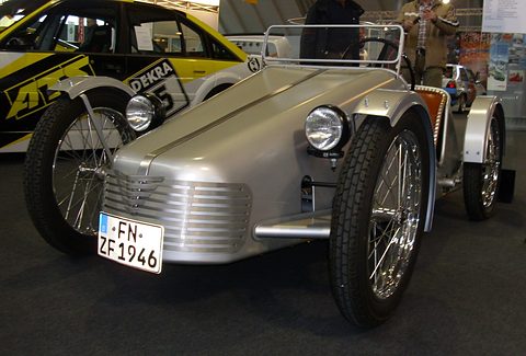 Silver doorless two-seater with a louvred nose and spoked wheels on a show stand.