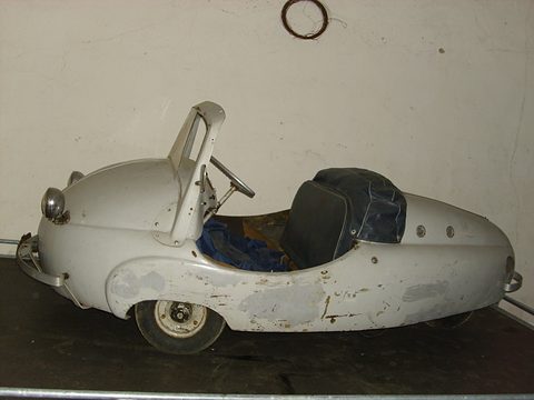 Open pale grey teardrop-shaped car in profile with patchy paint, a raised screen frame and a dark cover over the seat.