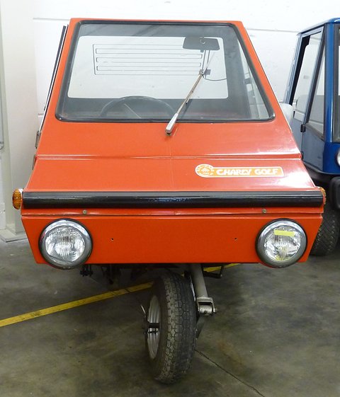 Front of an orange plastic three-wheeler with a tall flat windscreen, two round headlamps and one central front wheel.