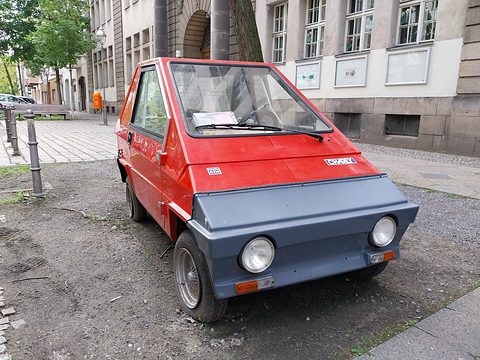 Small red plastic city car with a grey front panel, round headlamps and four wheels, parked on gravel in a street. Opens a larger view