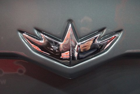 Chrome crown-shaped winged badge on dark grey paint, with red show lettering reflected in it. Opens a larger view