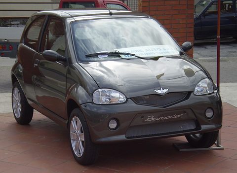 Dark grey small hatchback with a mesh grille, winged badge and a sign on its windscreen, on red tiled paving.