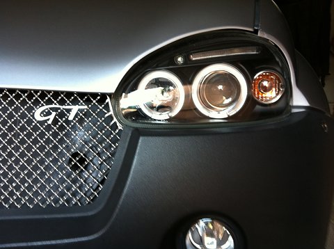 Close view of a black-framed headlamp with two ringed lenses beside a chrome mesh grille with GT lettering. Opens a larger view