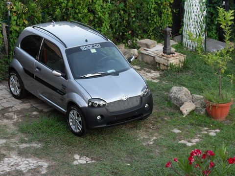 Silver small hatchback with black side stripes and a Sparco screen banner, seen from above on a grassy plot. Opens a larger view