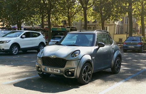 Matt grey small car with a black grille and alloy wheels in dappled shade, white SUVs and a bus behind. Opens a larger view