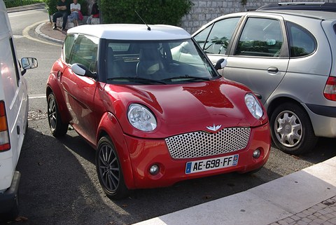 Red small two-door car with a white roof and perforated grille, parked between a white van and a silver MPV. Opens a larger view