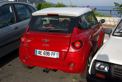 Rear of a red small car with a white roof and round tail lamps, squeezed between two parked cars, the sea behind. Opens a larger view