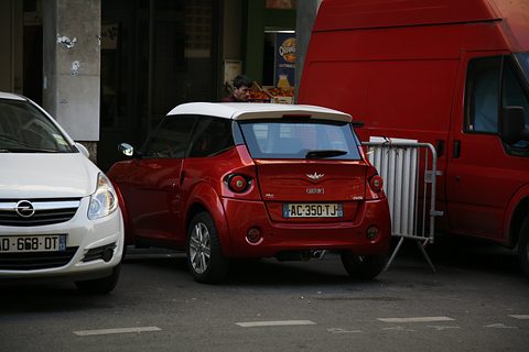 Rear of a small red two-door car with a white roof and winged badge, between a white hatchback and a red van. Opens a larger view