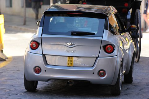 Rear of a silver small car with a winged badge, round tail lamps and a yellow plate, on cobbles. Opens a larger view