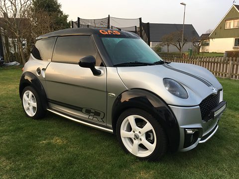 Silver-grey small car with black arch extensions, white wheels and side graphics, parked on grass by a wooden fence. Opens a larger view