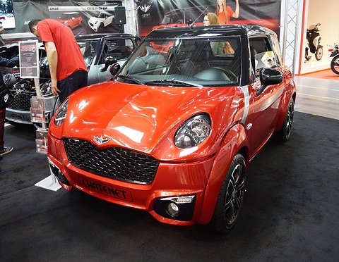 Bright orange small car with a black roof and honeycomb grille on a show stand, a man leaning in beside it. Opens a larger view