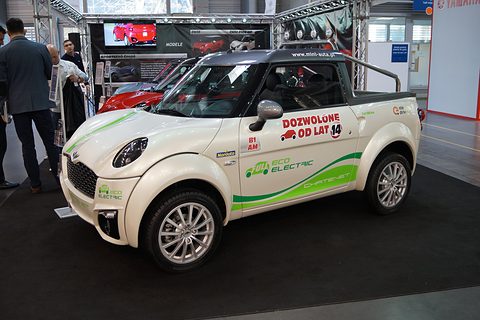 White small pick-up with green electric graphics and Polish dealer lettering on its door, on a show stand. Opens a larger view