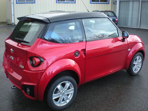 Glossy red small hatchback with a black roof and spoiler, seen from behind and to the left on wet tarmac. Opens a larger view