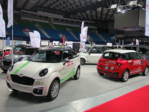 Two small Mini-like cars, one white with green electric graphics and one red, on a show stand in an arena. Opens a larger view