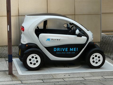 Side view of a white and black tandem two-seater with DRIVE ME! lettering in a marked bay by a wall. Opens a larger view