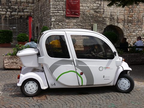 White four-seat electric car with doors and a green swoosh on cobbles in front of an old stone wall. Opens a larger view