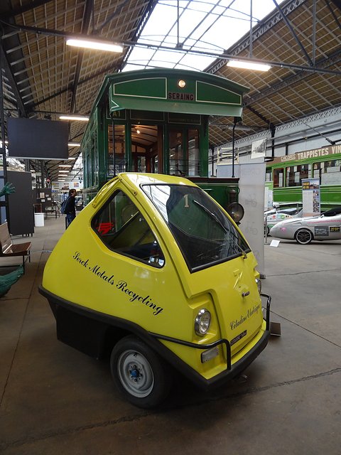 Yellow egg-shaped three-wheeler with script lettering on its side, parked in a tram depot with a green tram behind it.
