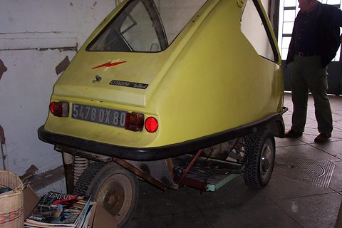 Yellow three-wheeler seen from the rear with plate 5478 DX 80, the battery cradle visible under the body, magazines on the floor.