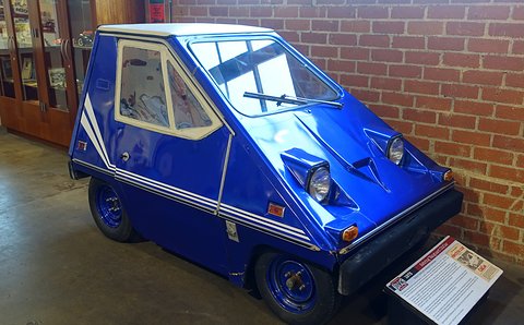 Glossy blue wedge-shaped electric car with white side stripes and a white roof, on a museum floor by a brick wall, an information placard in front. Opens a larger view