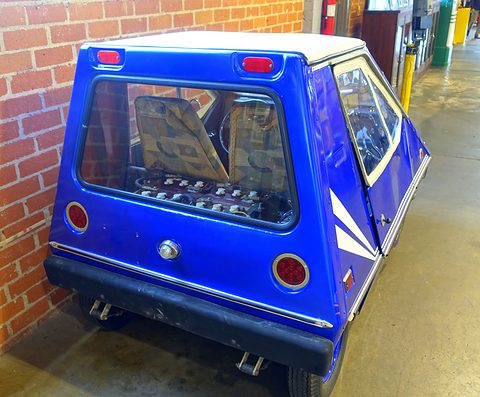Rear quarter of a blue wedge-shaped electric car with a large rear window, round red tail lamps and white stripes, beside a brick wall. Opens a larger view