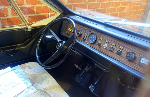 Interior of a small car: black steering wheel, woodgrain instrument panel with round gauges and a row of switches, patterned seat below.