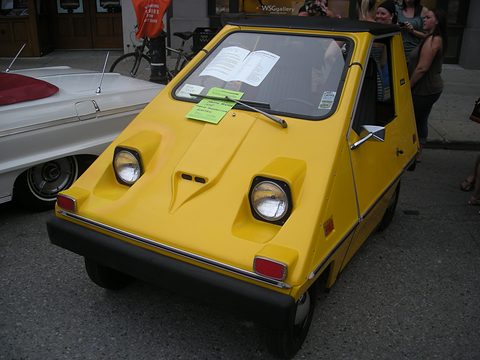 Head-on view of a yellow wedge-shaped electric car with two round headlamps, a black bumper and papers on the dashboard, a white classic car beside it. Opens a larger view
