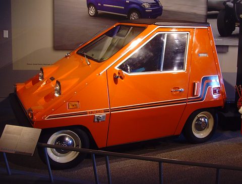 Orange wedge-shaped electric car with brown and white side stripes and whitewall tyres, on a museum floor behind a low rail.