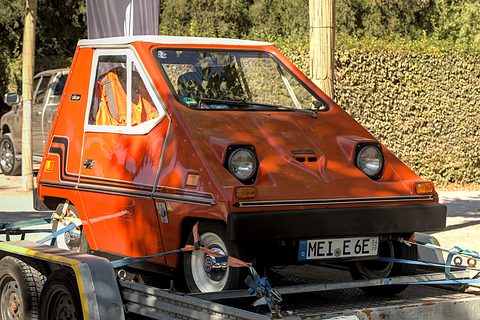 Orange wedge-shaped electric car with round headlamps and a German number plate, strapped onto a car trailer in front of a hedge. Opens a larger view
