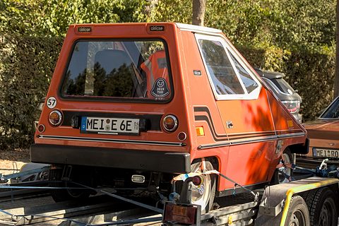 Rear of an orange wedge-shaped electric car on a trailer, with a large rear window, round tail lamps, side stripes and a German number plate. Opens a larger view