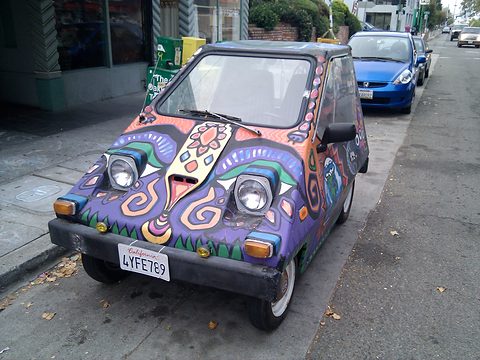 A small wedge-shaped car painted all over in bright purple, yellow and red patterns, parked on a city street. Opens a larger view
