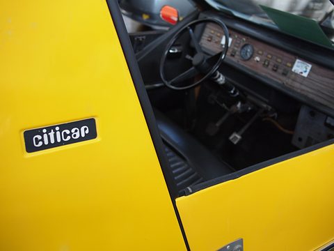 Close view of a yellow door with a black citicar script badge; beyond the door edge, a black steering wheel and a plain instrument panel. Opens a larger view