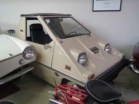 Cream wedge-shaped electric car with round headlamps and a folded black roof, parked between a white car and a red motorcycle indoors. Opens a larger view