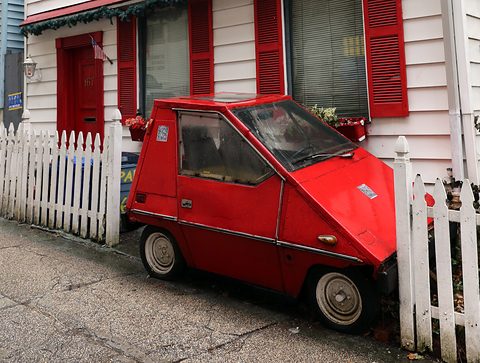 Small red wedge-shaped electric car with twin round headlamps, parked on a pavement beside a white picket fence and a white house with red shutters. Opens a larger view