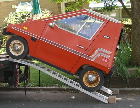 Red wedge-shaped electric car with white side stripes climbing a pair of metal ramps onto a trailer, trees and a building behind. Opens a larger view
