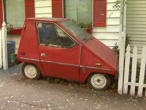 Weathered red wedge-shaped two-seater with a long sloping nose and slim chrome bumpers, beside a white fence and a house wall. Opens a larger view