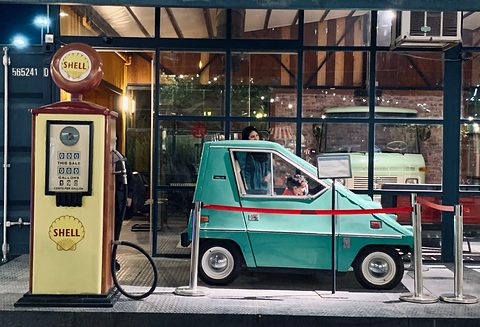 Pale teal wedge-shaped electric car inside a glass-fronted display at night, next to a cream Shell petrol pump with a round globe. Opens a larger view