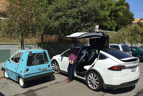 Small pale blue wedge-shaped car parked next to a white SUV with its rear doors raised like wings, a man standing between them.
