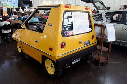Yellow wedge-shaped electric car seen from the rear quarter indoors, with notices taped to its rear window and an audience at a panel talk behind. Opens a larger view